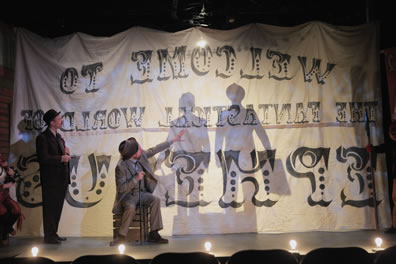 Solinus stands in dark suit and fedora holding a cigar in his hands to the left, Egeon sits on a crate, one hand on a cane between his legs, the other pointing up at the shadows of two men with suitcases through the carnival curtain. Stage footlights are visible across the front of the stage.
